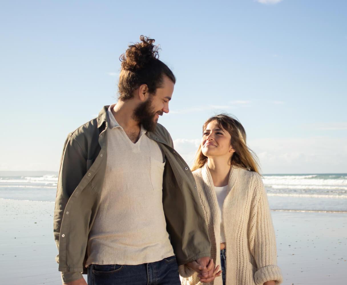 A man and woman walk together on the beach, their hands intertwined, with the ocean stretching out behind them.