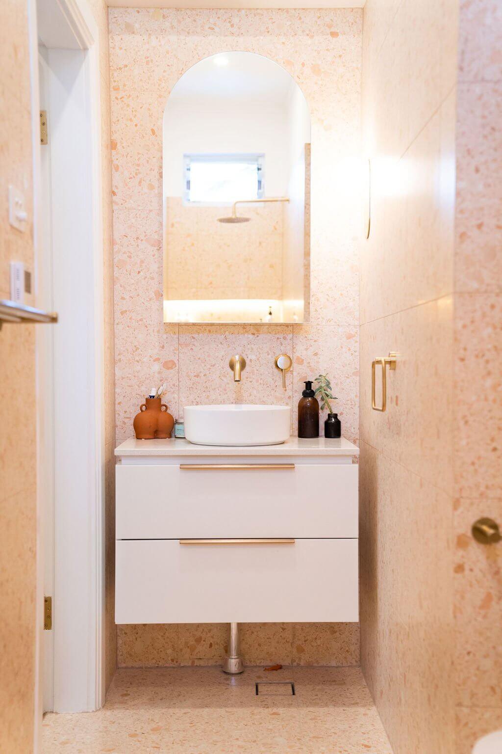 Contemporary bathroom with white vanity, round vessel sink, gold fixtures, and arched mirror on beige stone wall.