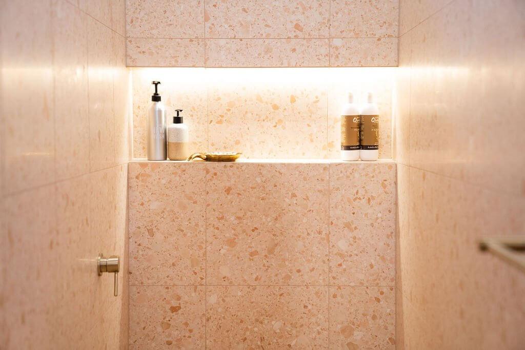 Peachy pink terrazzo tiled corner shelf styled with toiletries, warm lighting, Petersham bathroom renovation