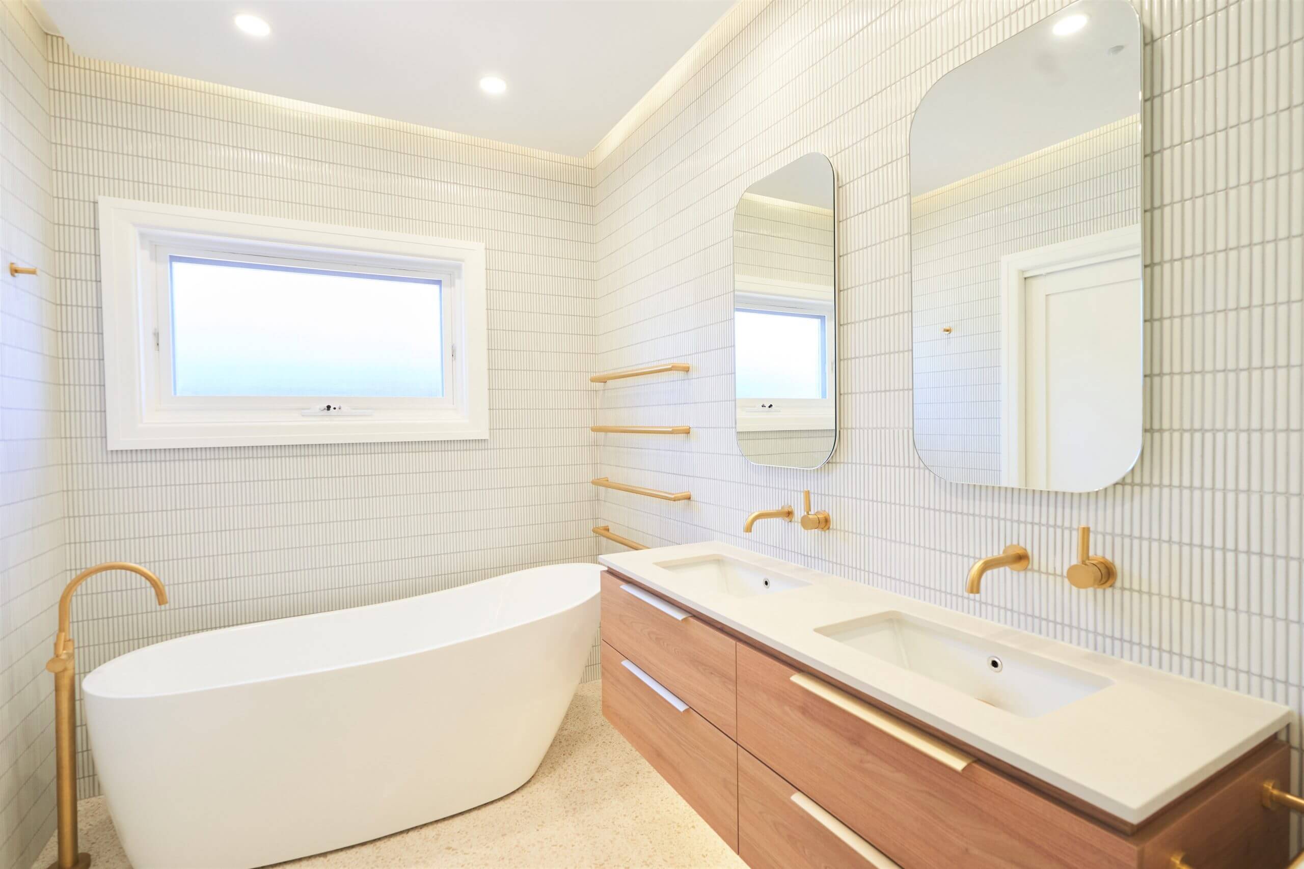 Luxury bathroom with freestanding white tub, double vanity with wooden cabinets, gold fixtures, and twin mirrors.