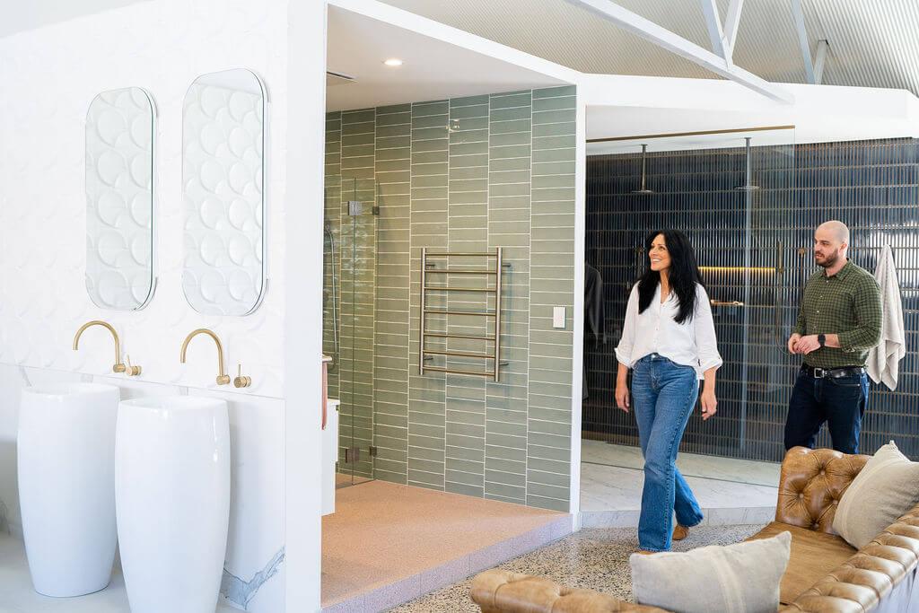 A man and woman walk in a bathroom that includes a shower, a mirror, and two basins with stylish golden taps.