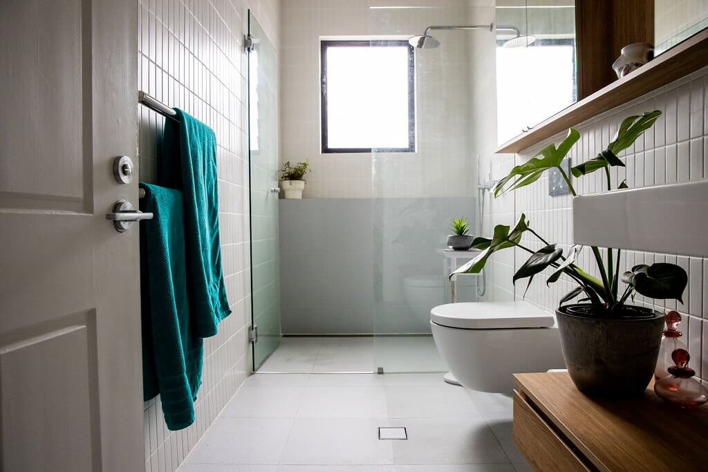 Modern bathroom featuring teal towels, a glass shower, potted plants, and minimalist decor with natural light from a window.