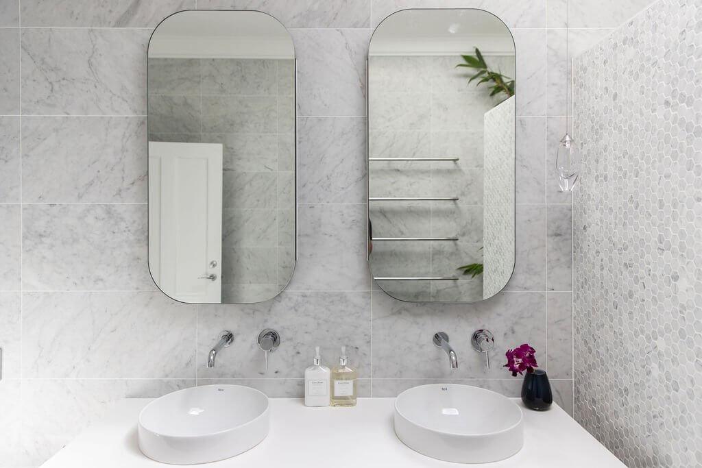 Elegant bathroom with dual vessel sinks, oval mirrors, marble wall tiles, and wall-mounted chrome tapware.