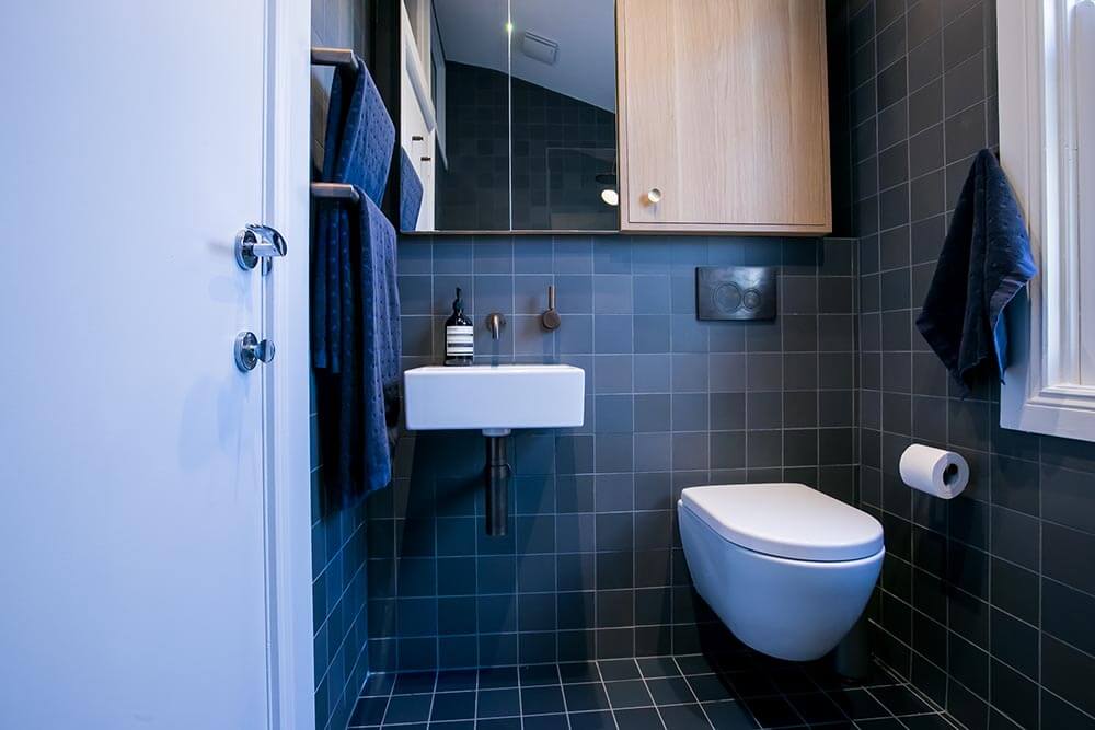 Modern bathroom with blue wall tiles, wall-mounted toilet, compact white basin, and sleek wooden cabinet