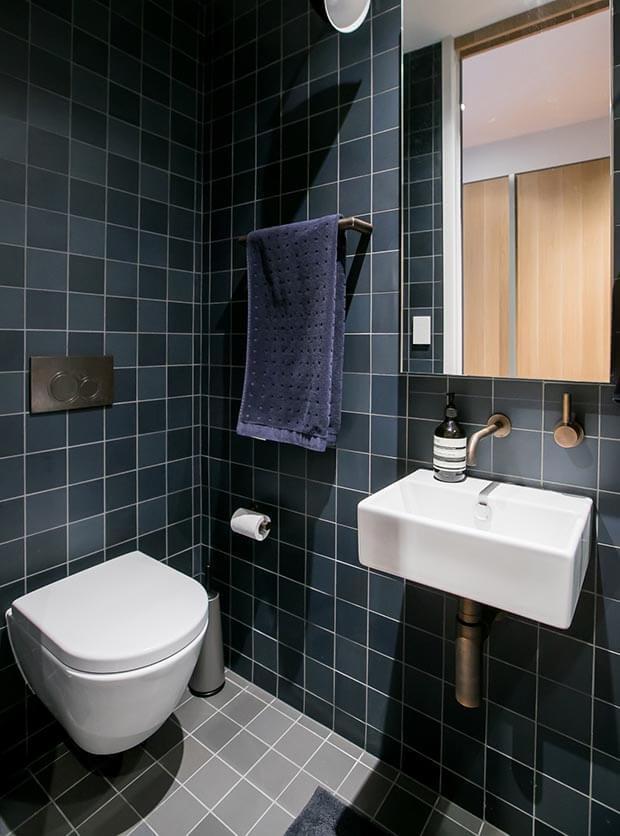 Modern bathroom with dark blue tiles, wall-mounted toilet, compact sink, and mirror for a sleek, space-saving design.