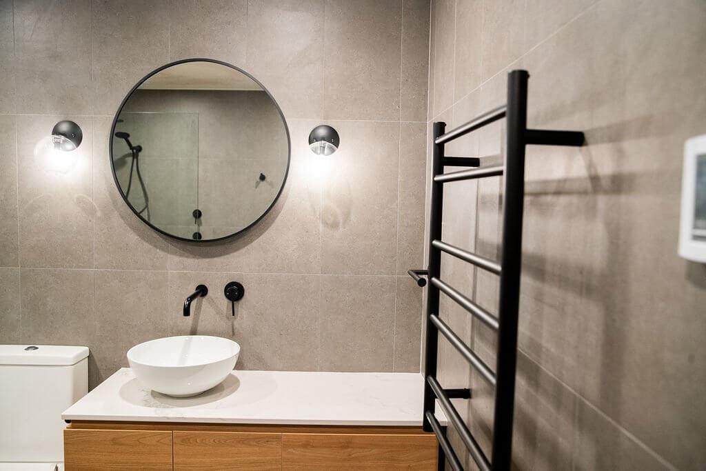Round mirror above timber vanity with vessel sink, matte black tapware and heated towel rail