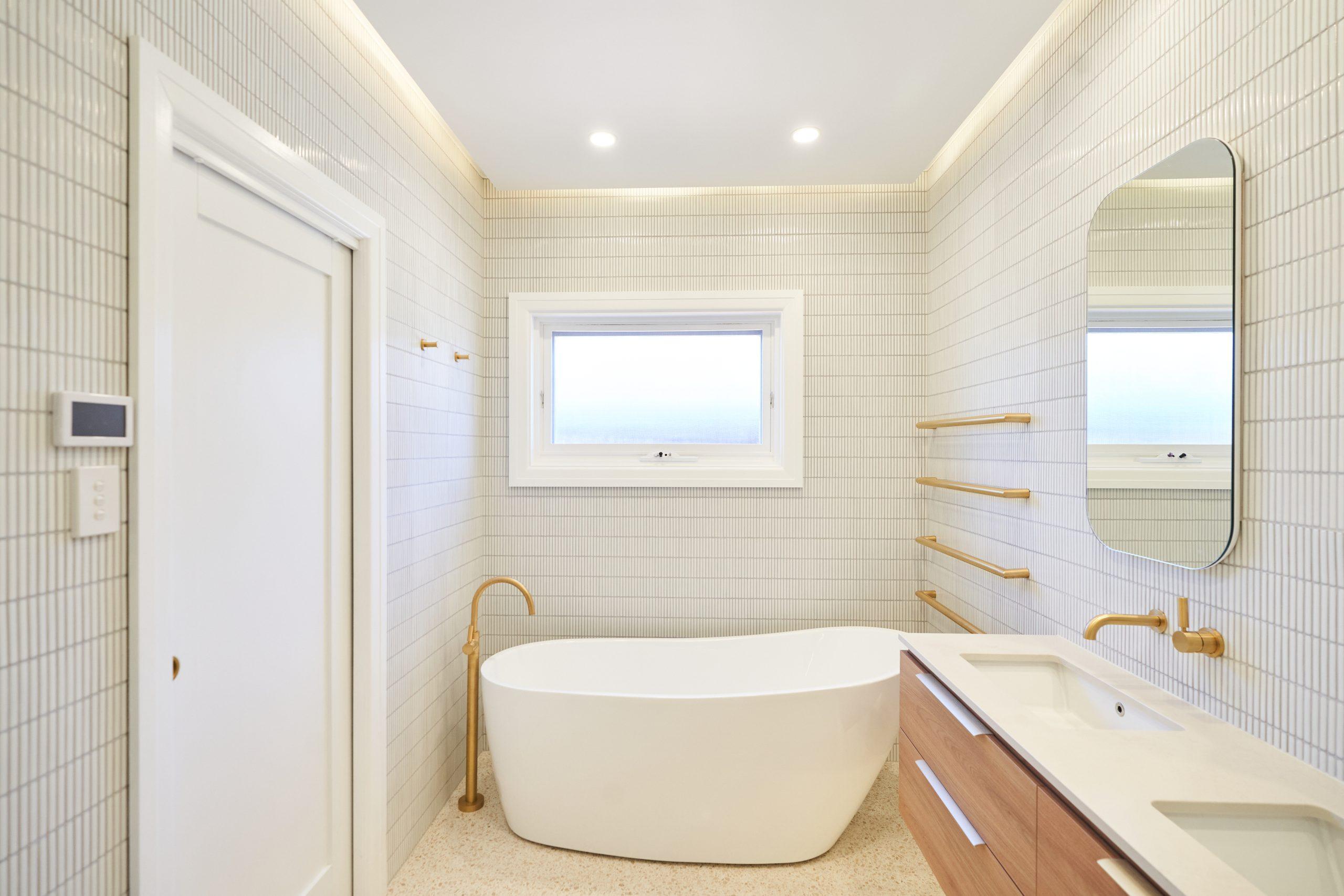 Modern ensuite with freestanding bathtub, terrazzo floor, vertical finger tiles, wood vanity, and brass tapware.