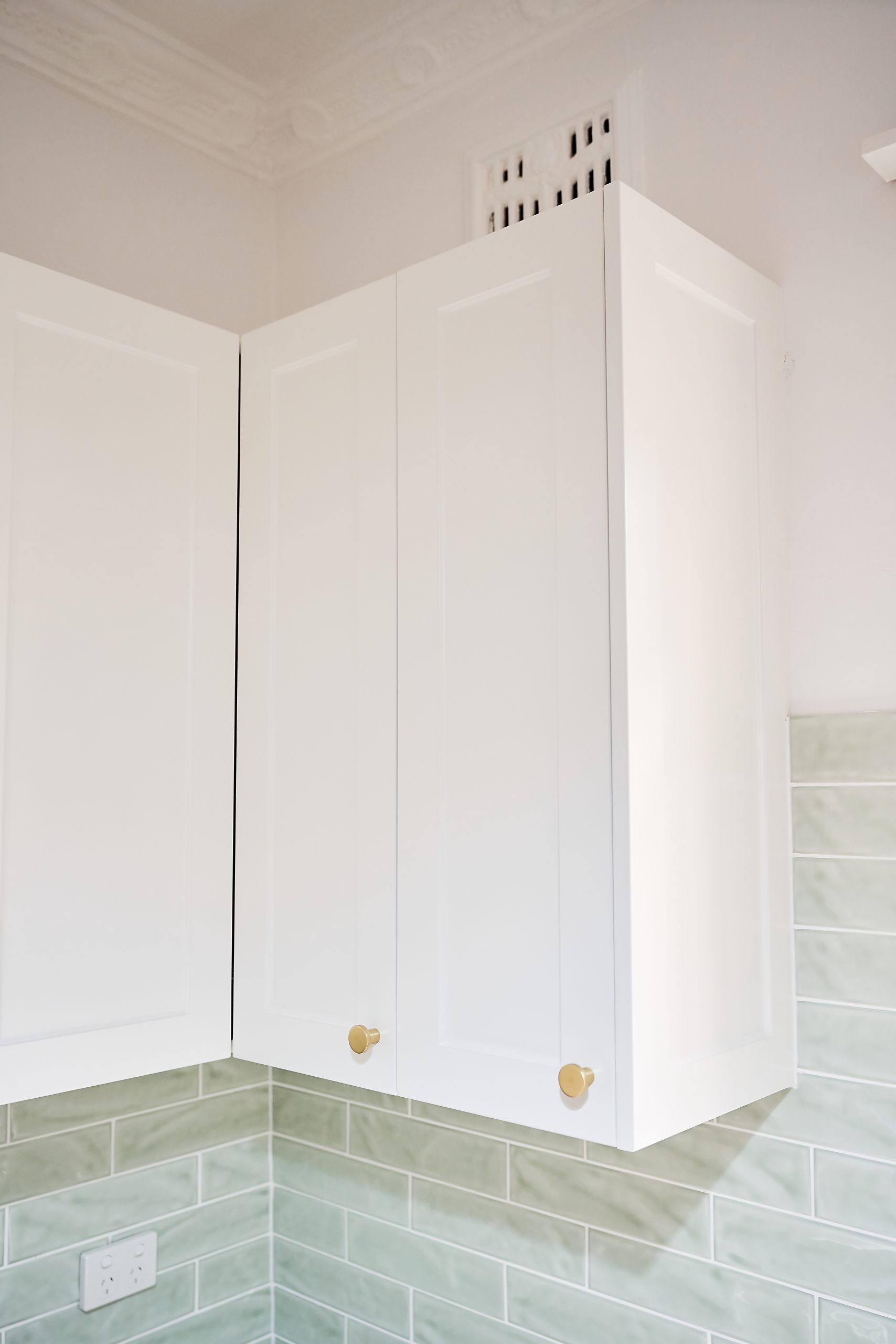 White overhead kitchen cabinet with golden knobs and green tiles splashback behind it