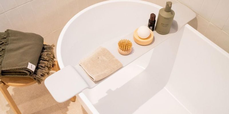 Modern white bathtub with bath caddy, towel, brush, and skincare products in a contemporary bathroom.